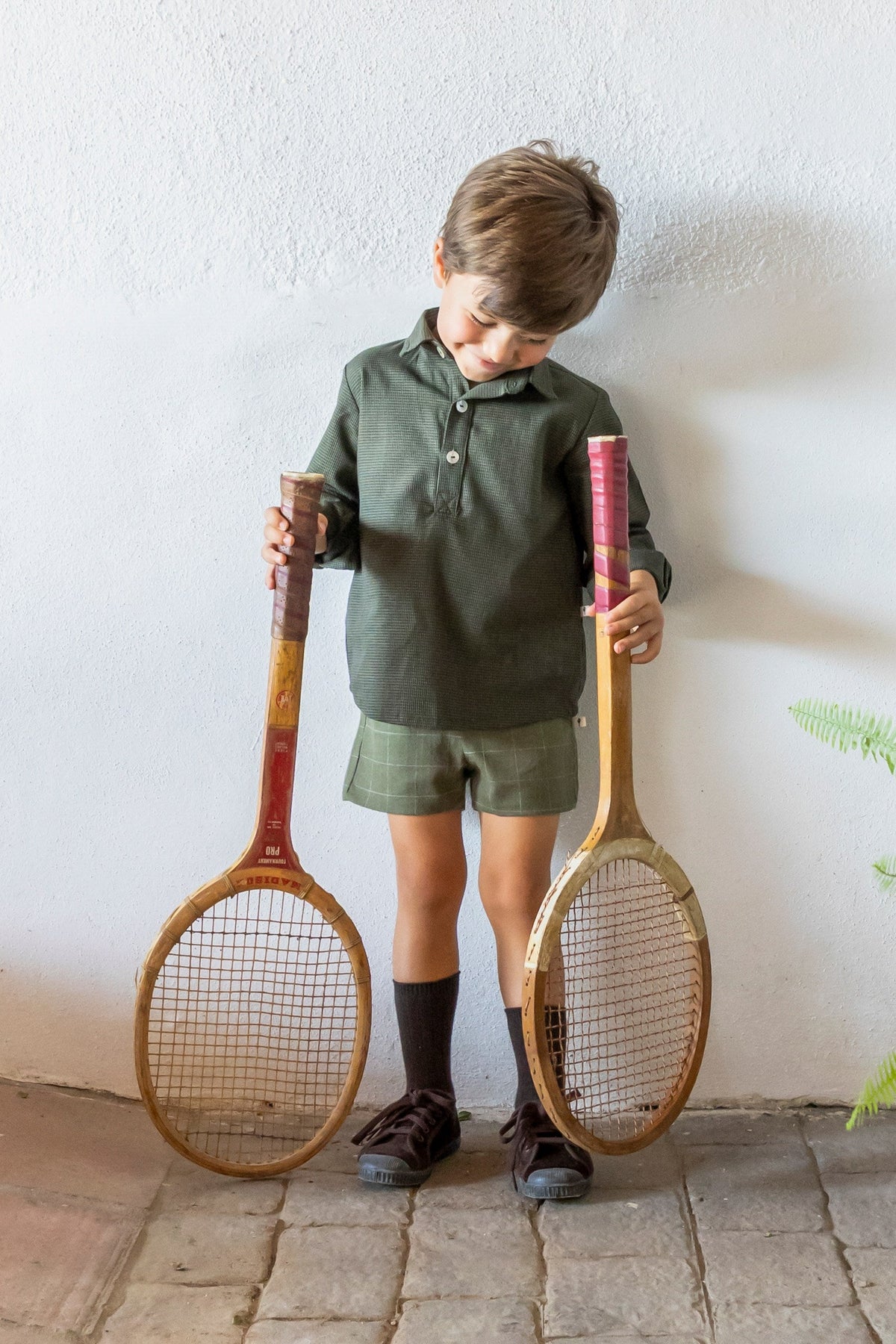 Niño con conjunto polo y short verde sosteniendo raquetas de tenis vintage